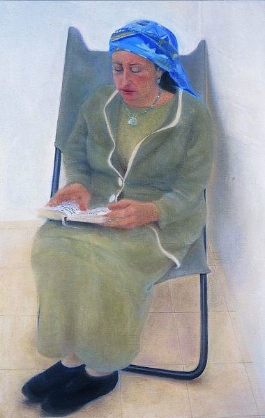 Woman looking down at book with Hebrew print on her lap, she wears a torquoise patterned scarf, sitting on grey chair.
